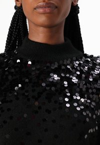 Maglione nero con collo alto, decorato con paillettes nere e argentate sovrapposte che creano un motivo testurizzato. Vista ravvicinata.