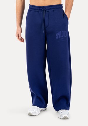 Mand iført løstsiddende marineblå joggingbukser med teksten "SMILODOX" på låret og hvide sportssko, står med den ene hånd i lommen.