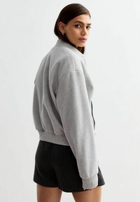 Szara krótka bluza z ribbowym kołnierzykiem i mankietami. Model nosi czarne spodenki w prążki, podkreślając luźny krój i fakturę odzieży.