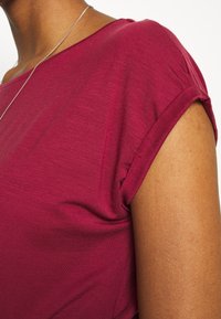 Gros plan d'une personne portant un haut rose foncé à manches courtes et un collier fin en argent sur fond blanc.