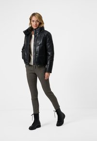 Svart läderpuffjacka med hög krage, beige stickad tröja under, bruna mönstrade slim-fit byxor och svarta snörskor.