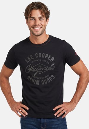 Homme souriant aux cheveux ondulés portant un t-shirt noir Lee Cooper avec un texte gris, les mains posées sur les hanches, sur un fond clair uni.