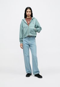 Νεαρή γυναίκα με μακριά σκούρα μαλλιά, που φοράει ανοιχτό τιρκουάζ φούτερ με φερμουάρ, καφέ μπλουζάκι, ανοιχτό μπλε τζιν και μαύρα αθλητικά παπούτσια, στέκεται σε λευκό φόντο.