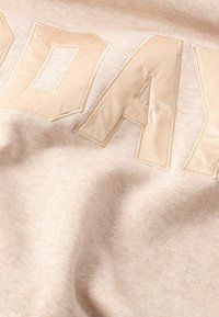 Tissu beige avec des lettres en relief et brillantes écrivant "AUJOURD'HUI". La texture est douce et légèrement élastique, avec une finition lisse et un minimum d'étirement.