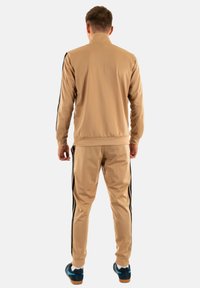 Uomo in piedi con le spalle voltate, vestito con una tuta beige con strisce nere sulle maniche e sui pantaloni, abbinata a scarpe sportive scure, su uno sfondo bianco.