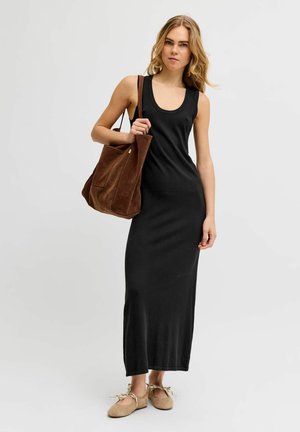 Femme portant une robe maxi noire sans manches, des ballerines beiges avec des nœuds, et portant un grand sac fourre-tout en suède marron sur l'épaule.