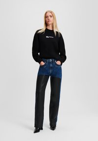 Črna kapuca s logotipom, modri kavbojke in črne usnjene hlače z mešano teksturo, gladka tekstura, široki nogi in črne koničaste čevlje.