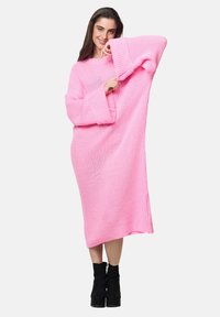 Übergroßes, rosafarbenes Strickkleid mit langen Ärmeln und Rippenmuster, ausgestattet mit lockerem Schnitt und mittellangem Design. Kombiniert mit schwarzen Stiefeln mit Absatz.