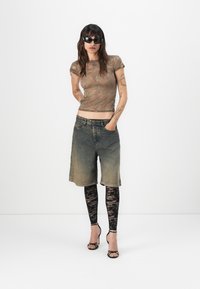 Top ajustado marrón con estampado, pantalones cortos de denim desgastados, leggins de encaje negros y tacones negros con tiras. La modelo lleva gafas de sol. Fondo minimalista.