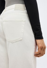 Κοντινό πλάνο ενός ατόμου που φοράει ανοιχτοκίτρινο τζιν με πίσω τσέπη και μπεζ λογότυπο, σε συνδυασμό με μαύρο μακρυμάνικο τοπ.