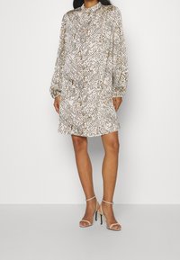 Robe à manches longues, de longueur genou en crème avec un motif abstrait noir et marron. Manches légèrement froncées et encolure ronde. Associée à des sandales à talons transparents.