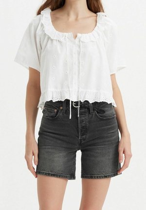 Femme portant un chemisier blanc à manches courtes avec des volants et des détails œillets, et un short en jean noir taille haute avec une ceinture noire.