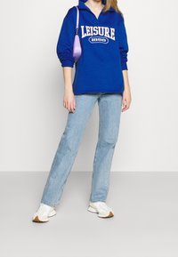 Blå fleece-tröja med texten "LEISURE 1998", kombinerad med ljusblå raka jeans och vita sneakers med krämfärgade detaljer.