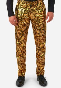 Zelta un melnu rakstu bikses ar zvaigžņu dizainiem un ģeometriskām līnijām; spīdīgs apdare; valkā ar melnām kurpēm un garām piedurknēm krekli.