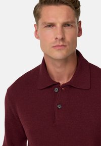 Bordeauxfarbiges Strickpolo mit klassischem Kragen, Knopfleiste mit drei Knöpfen und strukturiertem Stoff. Die Knöpfe sind dunkel mit einem subtilen Glanz.