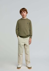 Olivgrüner Pullover mit Rundhalsausschnitt, beiges Cargo-Hose, weiße Sneakers mit grünen Akzenten. Das Model steht mit den Händen in den Taschen vor einem neutralen Hintergrund.