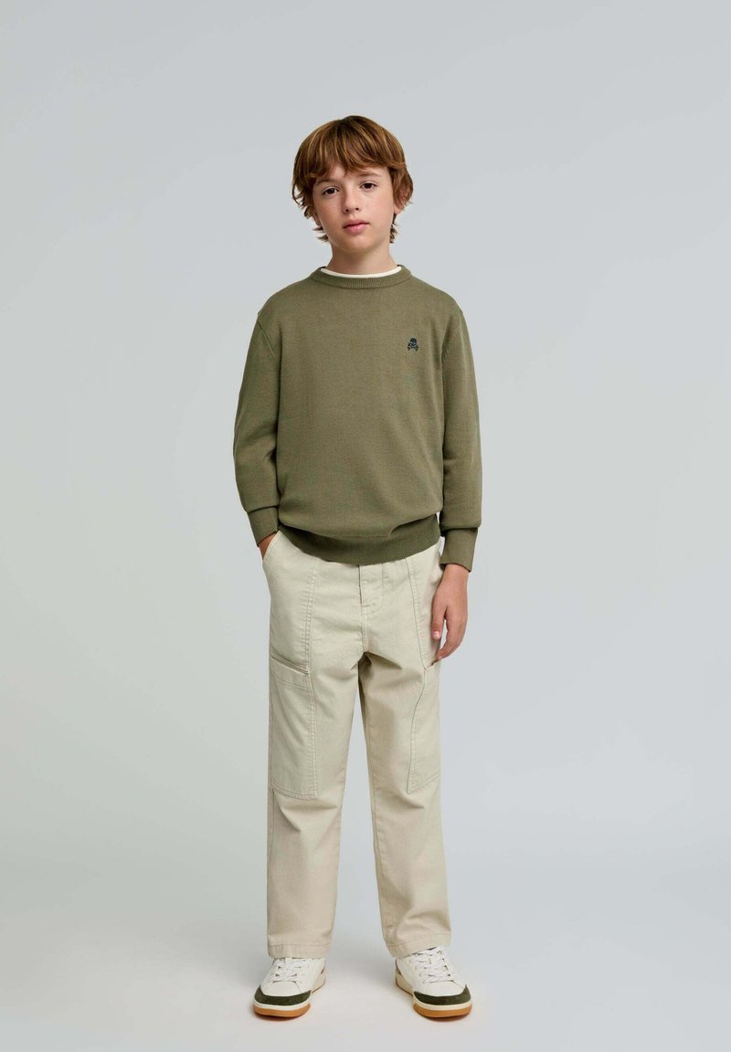 Olivgrüner Pullover mit Rundhalsausschnitt, beiges Cargo-Hose, weiße Sneakers mit grünen Akzenten. Das Model steht mit den Händen in den Taschen vor einem neutralen Hintergrund.