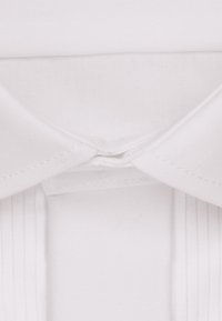 Dettaglio del colletto di una camicia bianca con una texture liscia, forma strutturata e tessuto plissettato nella spalla, caratterizzato da cuciture ordinate.