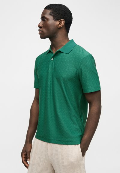 Άνδρας που φοράει ένα υφασμένο σκοτεινό πράσινο κοντομάνικο πόλο και ανοιχτό μπεζ παντελόνι, στέκεται με το ένα χέρι στην τσέπη, κοιτάζοντας προς τα αριστερά.