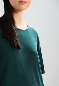 T-shirt en tissu doux de couleur vert foncé, avec un col rond et des manches courtes, doté d'une coupe décontractée et d'une texture lisse.