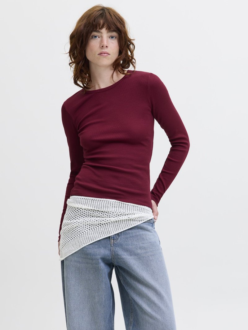 Tânără cu păr ondulat și castaniu, purtând un top bordo cu mâneci lungi mulat, blugi deschiși la culoare și un cardigan alb legat în jurul taliei.