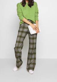 Groene trui met structuur, grijze geruite broek met gele accenten en witte sneakers. Model houdt een kleine witte clutch vast.