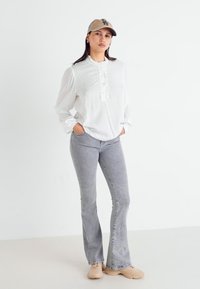 Camicia bianca con bottoni e pois testurizzati, jeans grigi a zampa d'elefante, scarpe beige e un berretto marrone chiaro con logo, in piedi contro uno sfondo neutro.