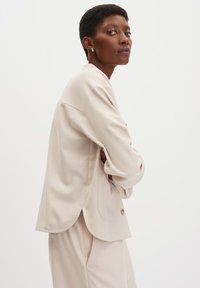 Licht beige longsleeve shirt met een losse pasvorm, ronde zoom en zijsplitten. Heeft een enkele knoopaccent en een zachte textuur.