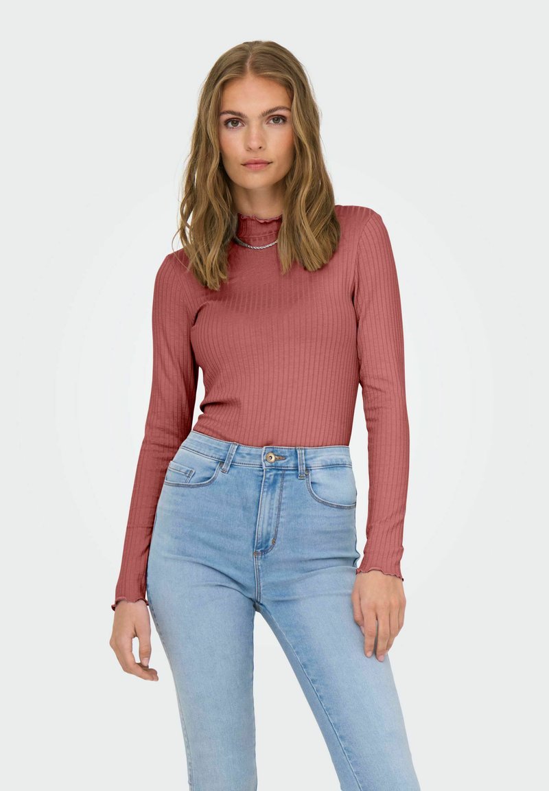 Top a maniche lunghe a coste in rosa polveroso con colletto alto e arricciato. Abbinato a jeans in denim blu a vita alta. Texture liscia.