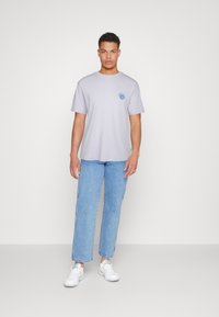 Tricou gri deschis cu mânecă scurtă, cu un grafic de soare pe pieptul stâng, asortat cu blugi drepți de un albastru deschis și adidași albi.