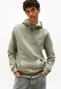 Giovane uomo che indossa una felpa verde salvia con logo viola "Tommy Jeans", con le mani nelle tasche, sovrapposta a una maglietta a righe e jeans di denim chiaro.