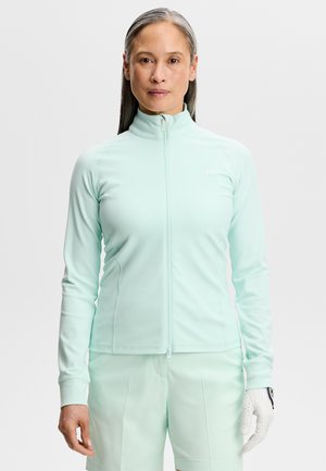 Midaldrende kvinde med glat gråt hår iført let mintfarvet lynlåstrøje og matchende shorts, med hvid golfhandske på højre hånd.