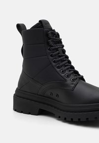 Botte de combat noire à lacets avec cheville rembourrée, semelle épaisse et languette à boucle à l’arrière, présentée sur un fond uni clair.