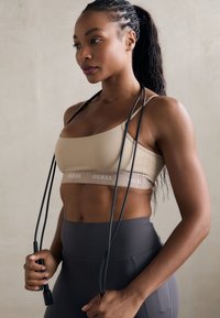 Sujetador deportivo beige con banda con logo, combinado con leggings oscuros de cintura alta. Cuerda de saltar oscura sostenida en las manos de la modelo, con textura suave.