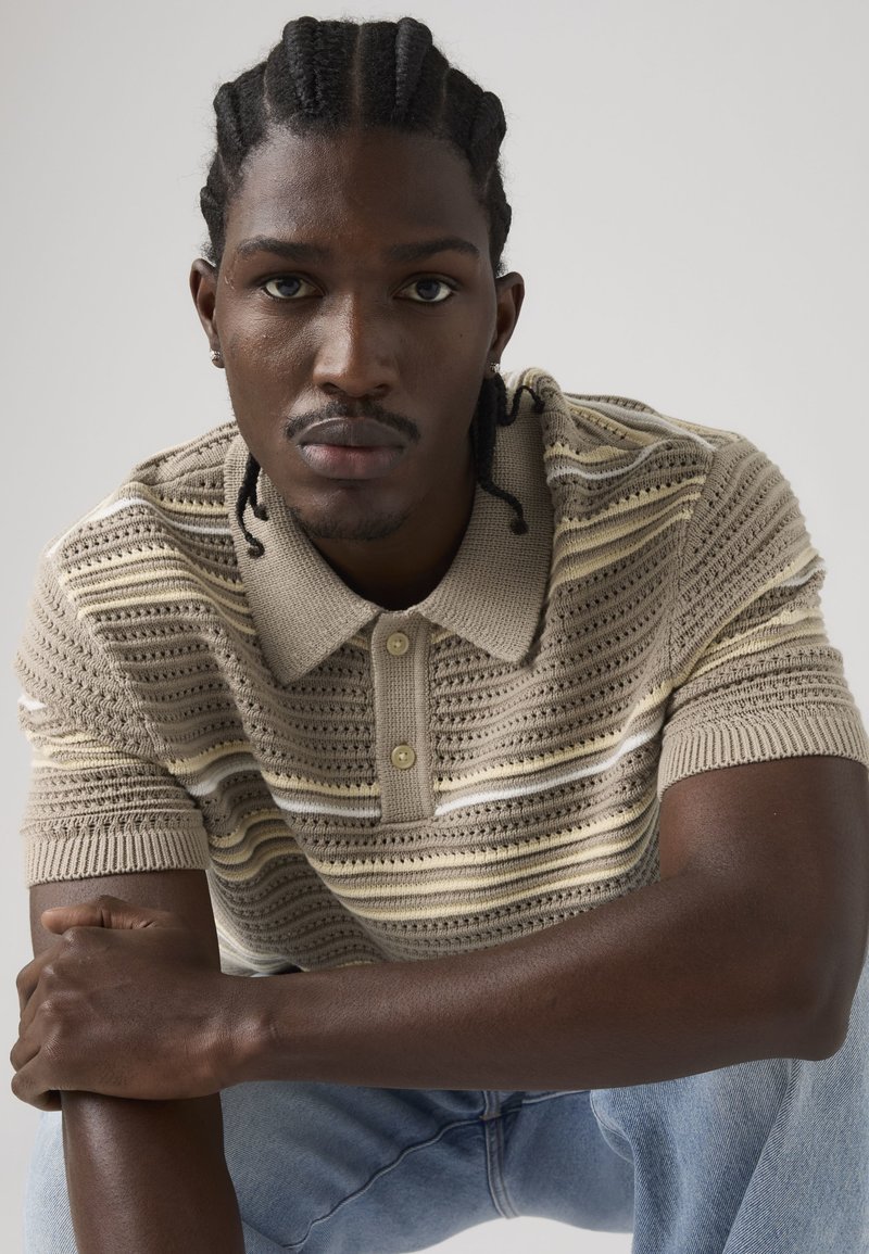 Homme aux cheveux tressés portant un polo en maille beige rayé et un jean bleu clair, assis les bras croisés et regardant droit devant.