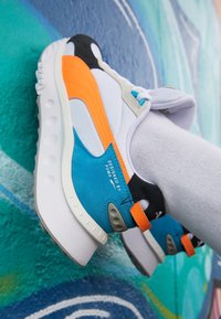 Sneaker multicolori con dettagli bianchi, neri, arancioni e blu, caratterizzati da una suola texturizzata e un'etichetta "DESIGNED BY PUMA" sul lato.