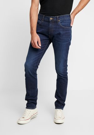 Homme portant un jean slim bleu foncé et des baskets blanches, debout devant un fond uni avec une main dans la poche.