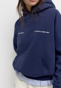 Marineblauwe hoodie met een voorzak, voorzien van witte tekst "WALKING PROUD" en "AUTHENTIC VIBES ONLY." Gecombineerd met een gestreepte zoom.