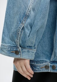 Gros plan d'une main reposant sur le côté, portant une veste en jean bleu clair avec des coutures visibles et des boutons métalliques.