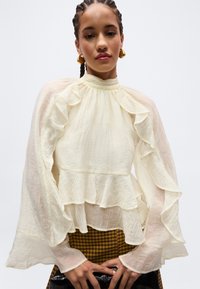 Γυναίκα με πλεξούδες φοράει διαφανές ελεφαντόδοντο πουκάμισο με βολάν και κρατάει μαύρο τσαντάκι με υφή σε απλό φόντο.