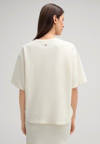 Cremfarbens, oversized T-Shirt aus Baumwolle mit kurzen Ä sleeves, abgerundetem Saum, rohen Säumen und einem kleinen metallischen Akzent am Nacken hinten.