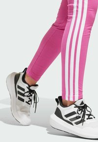 Leggings sportivi rosa con strisce bianche sui lati abbinati a scarpe da ginnastica nere e bianche con una parte superiore testurizzata e lacci prominent.