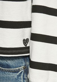 Maglia a maniche lunghe a righe bianche e nere con tessuto testurizzato, caratterizzata da un logo a forma di cuore nell'angolo in basso, abbinata a jeans blu.