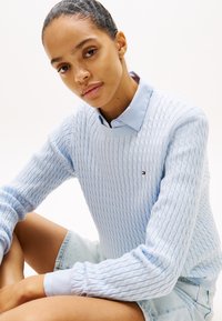 Lyseblå kabelstrikket genser laget av mykt materiale, med ribbestrikket hals og subtil logo. Brukt over en skjortekrage.