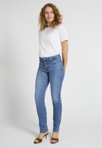 Lokkis juustega naine, kes kannab valget t-särki, siniseid kõrge vöökohaga kitsaid teksapükse ja pruune avatud varbaga kontsakingi, seisab valge tausta ees.