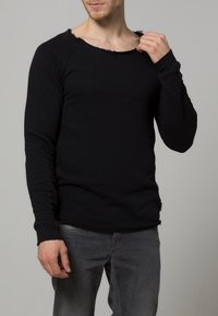 Chemise noire à manches longues avec un col rond, présentant des bords effilochés et une coupe décontractée. Fabriquée en tissu doux, elle est assortie à un jean gris.