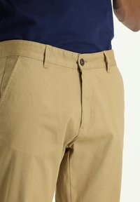 Chinos khaki con vestibilità slim, materiale in cotone, vita standard e chiusura con bottone in metallo. Parte anteriore piatta con tasche laterali.
