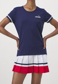 Mörkblå kortärmad träningsskjorta med vit trim och logotyp, tillsammans med en plisserad kjol med röda, vita och mörkblå ränder.