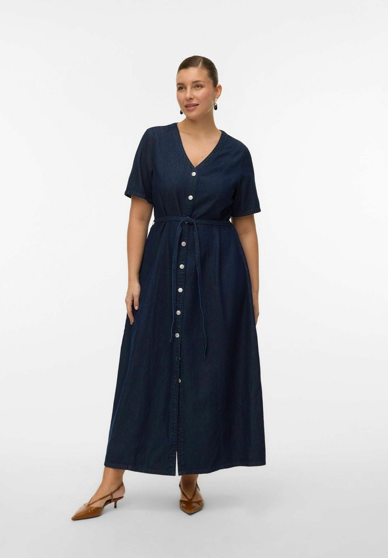 Mujer con un vestido midi de mezclilla azul oscuro abotonado, de mangas cortas y cintura anudada, combinado con tacones marrones puntiagudos con tira trasera.