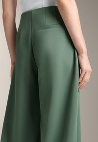 Zelene široke hlače iz teksturirane tkanine, z zadnjim žepom in prilagojenim krojem, prikazane s strani z roko, naslonjeno na bok.